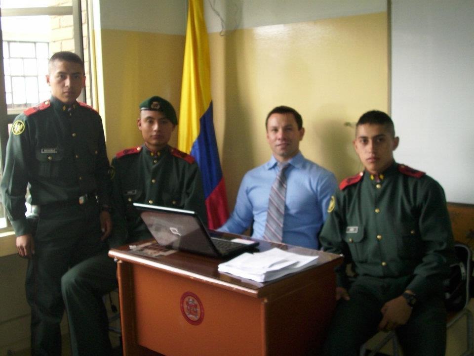 John seated at a classroom desk with uniformed students and the Colombian flag.